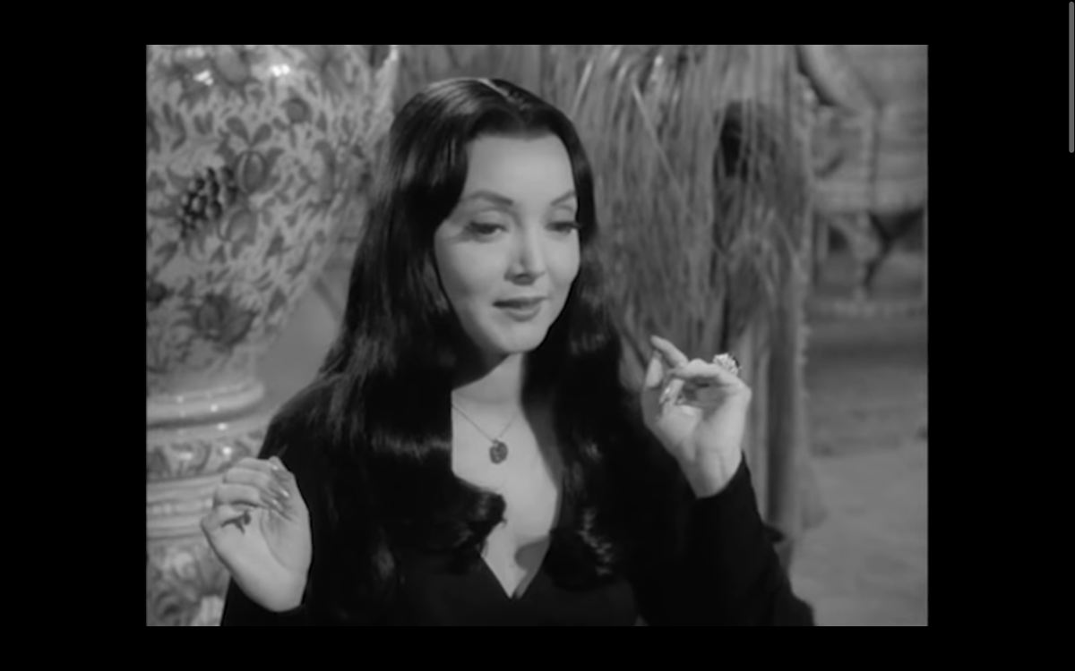 Morticia Addams wearing black. Her hands are by her shoulders.
